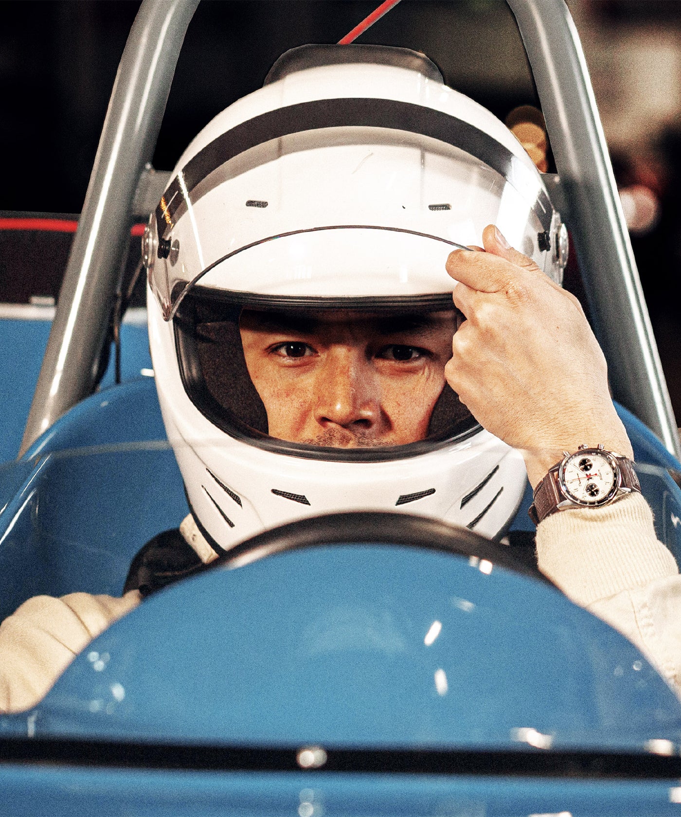 Un pilote de formule 1 des années 60 au volant qui ferme la visière de son casque avec sa montre ronde allure legend 60s panda au poignet